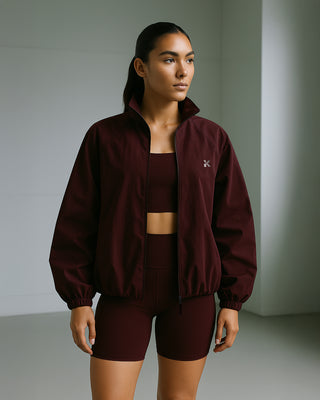 Athletic woman in a minimalist indoor studio wearing a matching Kinia set in deep plum. She sports a structured, lightweight zip-up jacket with a bubble hem and raglan sleeves, paired with sculpting high-waisted performance shorts and a coordinating sports bra. The technical fabric has a soft, matte finish with subtle volume, creating a sleek and functional silhouette ideal for training or elevated athleisure wear.