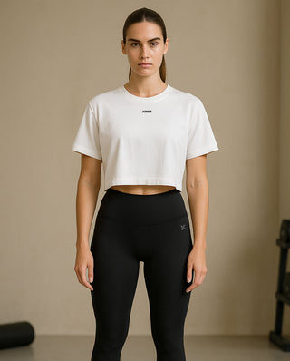 Fit woman wearing a cropped white Kinia t-shirt and high-waisted black leggings, posing in a soft-lit gym space with minimalist fitness equipment behind her.